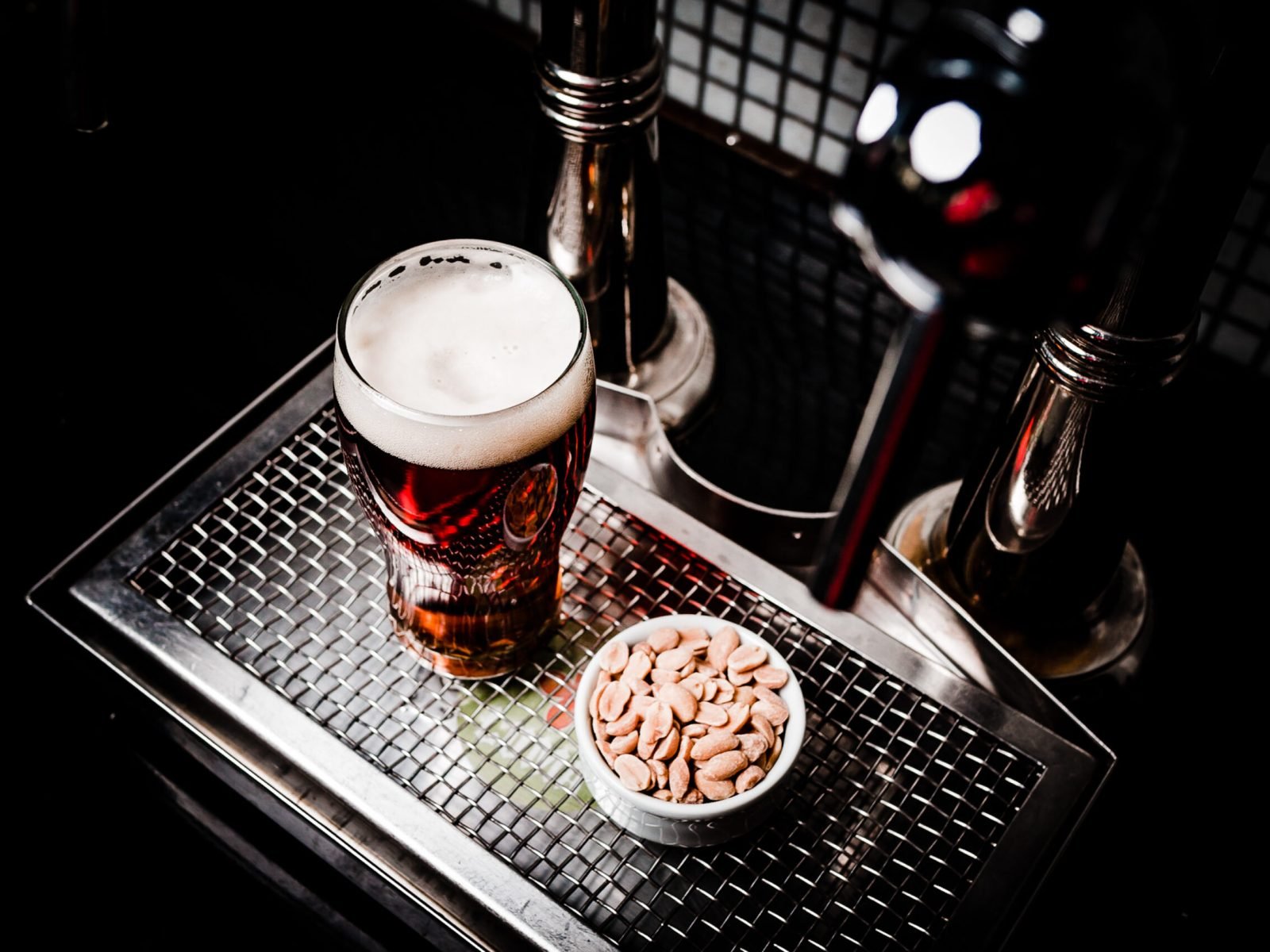 A top view shot of a glass of lager beer with a cup of peanuts on a metal screen tray